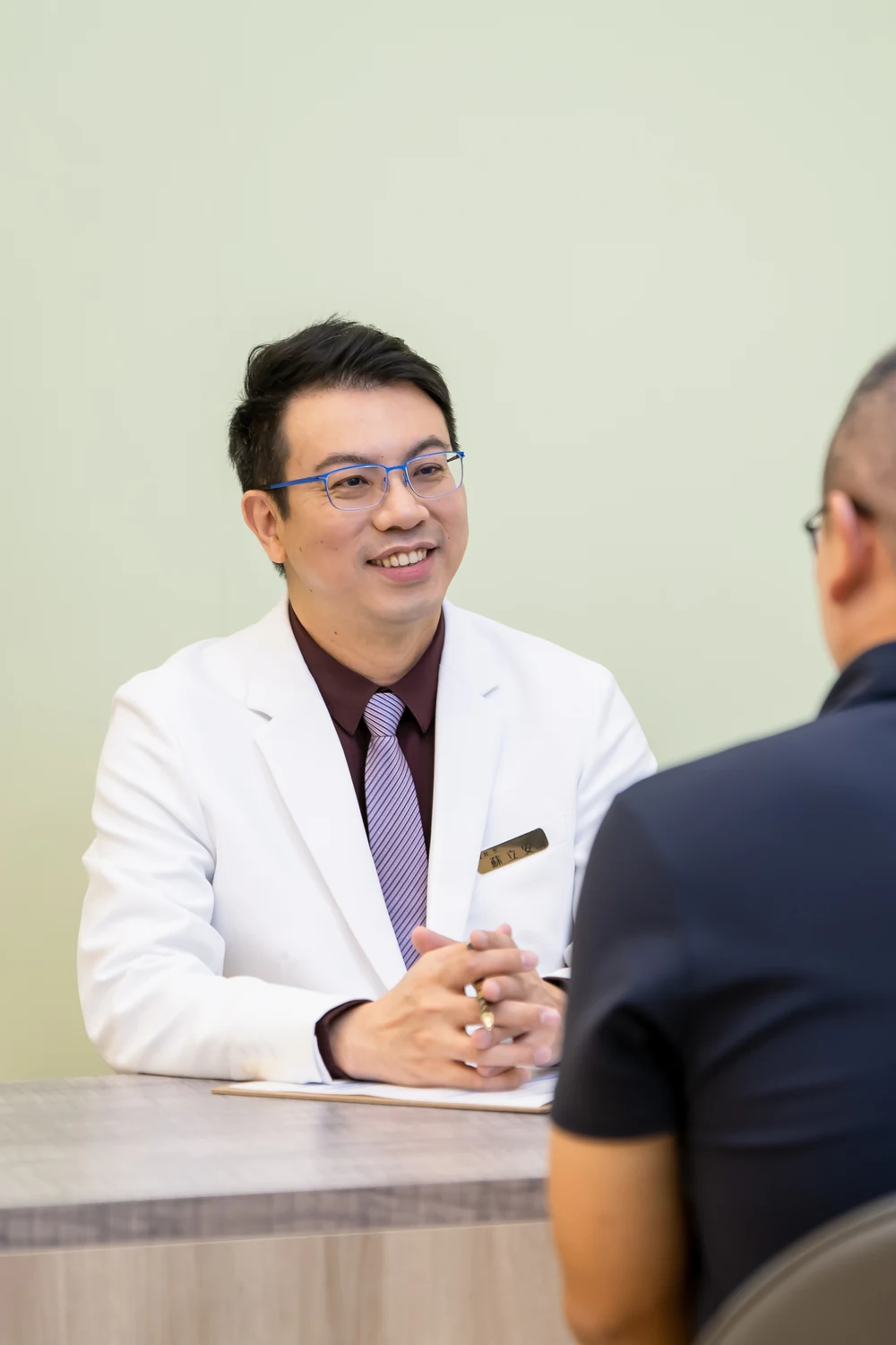 Dr. Tony Su in a physician's white coat, mid-consultation with a patient, warm and attentive