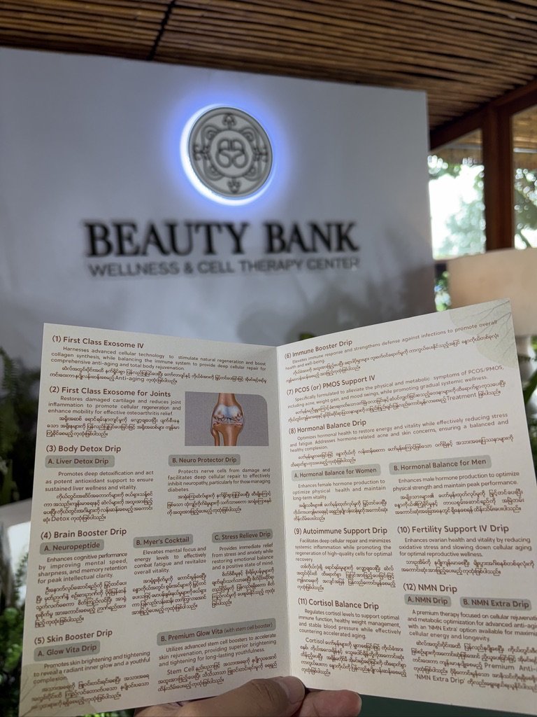 The bilingual English and Burmese IV Drip menu at Beauty Bank — from First Class Exosome IV and Brain Booster to Hormonal Balance and NMN — held up in front of the illuminated clinic sign.