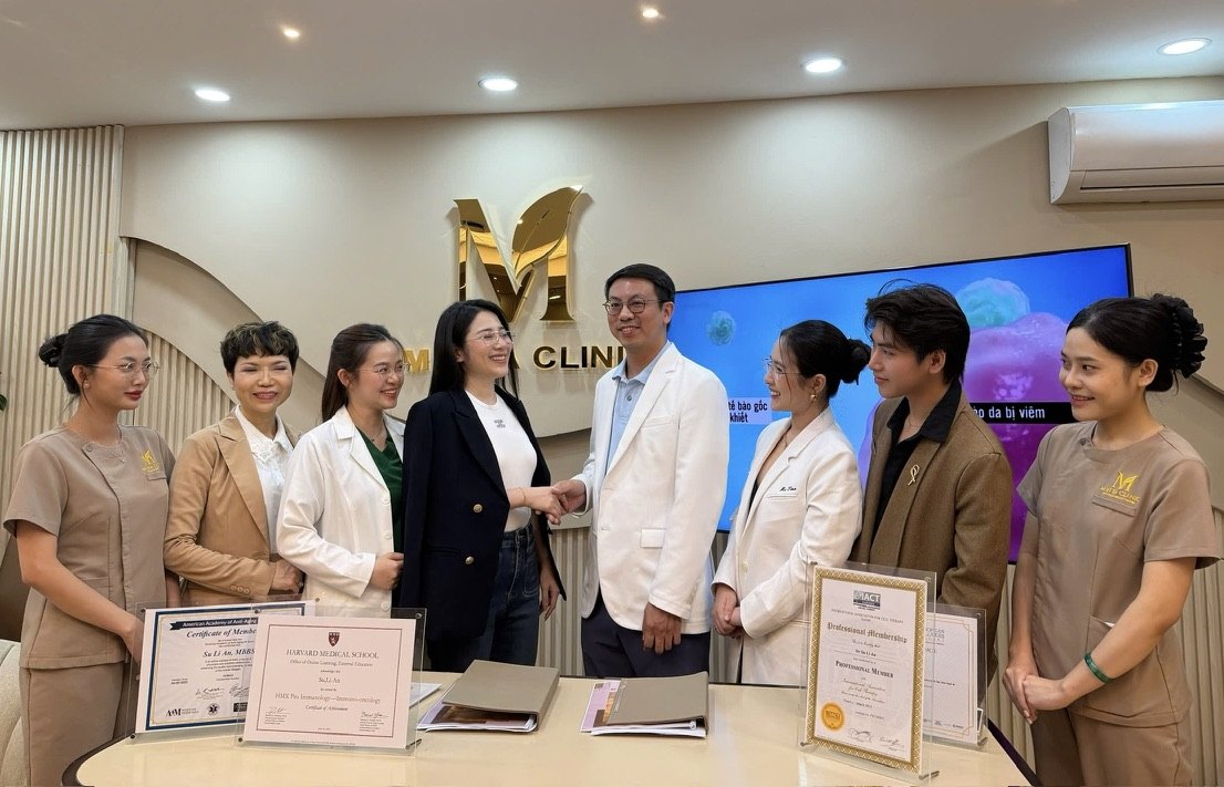 Dr. Tony Su shaking hands with the M VITA founder in the M VITA consultation room in front of the clinic team, formalising the R2-IWAA · M VITA cooperation.